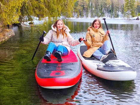 Kirov, Russia - March 14, 2024: Two women enjoy paddle boarding on a serene lake during winter, surrounded by lush trees and a tranquil atmosphere.