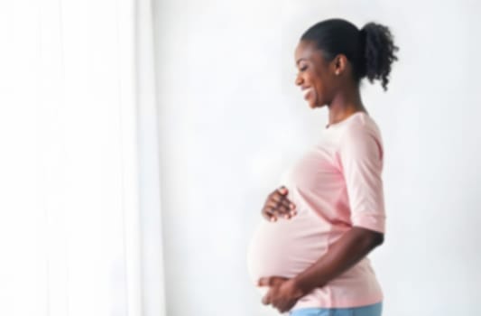 Happy expectant mother stands by a window, cradling her baby bump with a joyful expression in a softly lit room.. Blurred picture