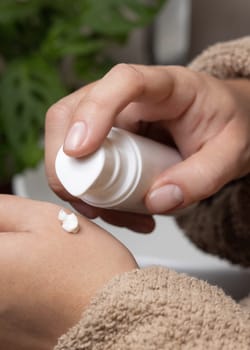 Close up of woman in brown bathrobe applying moisturizing cream on hand and holding a cosmetic bottle in other hand in bathroom. Body and hand care concept