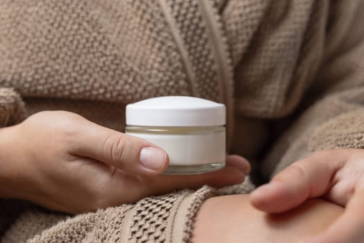 Close up of woman in light brown bathrobe holding white glass cream jar in hands with natural manicure, mockup. Facecare concept. Cropped Image of cosmetic blank container