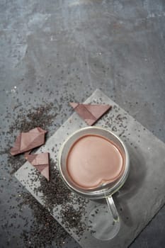 Glass cup of hot chocolate in heart shape on table surrounded by chocolate pieces and black chia seeds on grey textured background, Desert concept in color of the year 2025, mocha mousse,