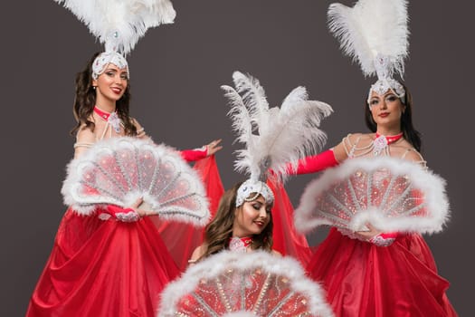 Three performers in red gowns and feathered headpieces gracefully present a captivating dance with ornate fans at an event.