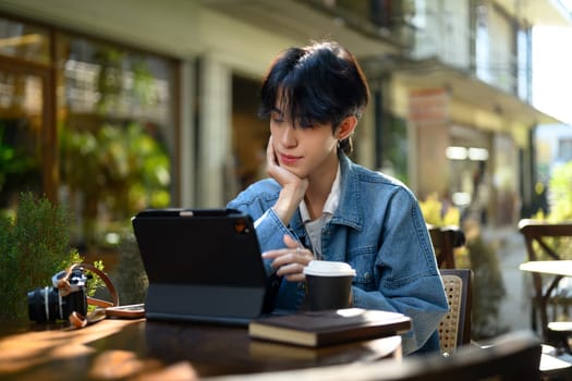 Stylish young man wearing a denim jacket using digital tablet at an outdoor cafe.