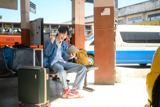 Excited traveler looking at digital tablet and celebrating sitting in a transportation terminal with a suitcase.