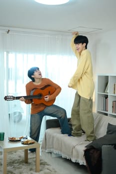 Cheerful gay couple enjoying casual moment at home playing the guitar and singing in living room.
