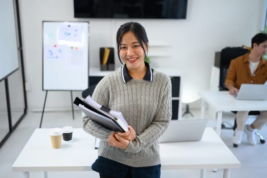 Portrait of smiling young woman standing in a bright office holding stack of documents and binders.