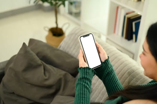 Close up of young woman sitting on a couch holding a smartphone with a blank white screen.