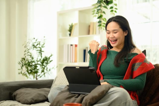 Happy woman in a green sweater celebrating while using digital tablet on her lap.