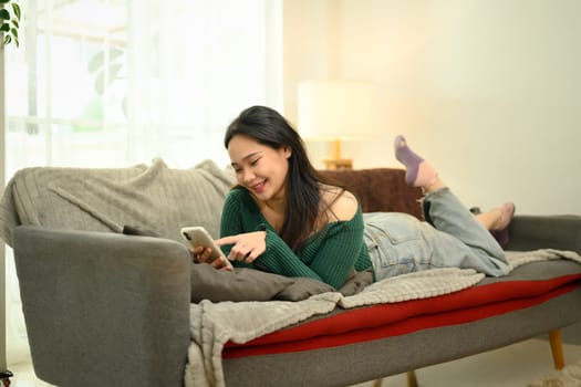 Relaxed asian woman reclining on a cozy couch and using mobile phone.