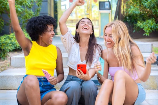 Three joyful young women sit on outdoor steps, excitedly celebrating while looking at a smartphone. Their cheerful expressions and casual summer outfits create a lively and friendly urban atmosphere.