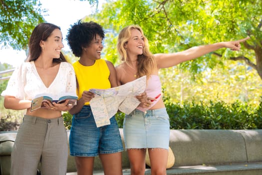 Three cheerful young women stand outdoors, holding maps and a travel guide while pointing at something. They smile excitedly, enjoying their adventure in a sunny urban setting with trees and buildings