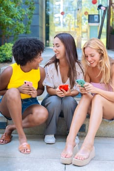 Three happy young women sit on outdoor steps, each engaged with their smartphone. They smile while looking at their screens, dressed in casual summer outfits, creating a relaxed and social atmosphere.