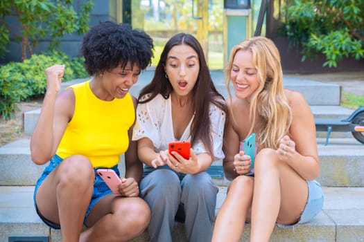 Three joyful young women sit on outdoor steps, excitedly celebrating while looking at a smartphone. Their cheerful expressions and casual summer outfits create a lively and friendly urban atmosphere.