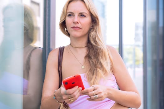A blonde woman standing outdoors, focused on her smartphone. Captured in a modern urban environment, highlighting casual elegance, digital connectivity, and a relaxed lifestyle.