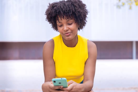 A young woman with natural curly hair wearing a yellow top rests her face on her hand while holding a smartphone. Calm and happy moment captured in a bright outdoor setting.
