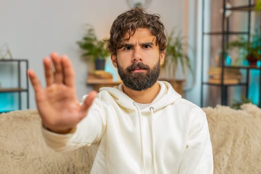 Portrait of Indian young man guy on sofa couch with serious expression doing stop gesture with hand at home, warning of finish, prohibited access, declining communication, and body language danger.