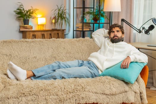 Tired Indian handsome young man enjoy relaxing on home sofa in living room. Happy guy lying on couch resting napping after hard working day, closed his eyes, put his hand behind his head, stress free.
