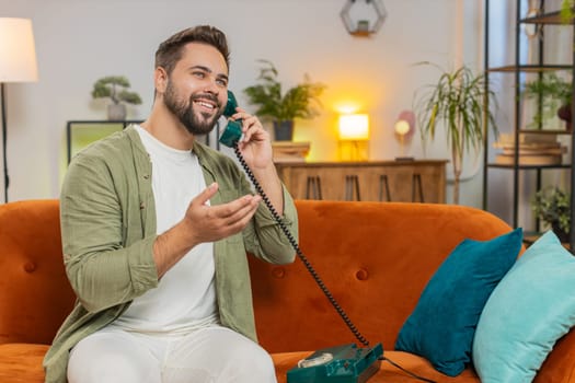 Smiling young man making wired telephone call conversation with friends sitting on couch at home. Happy excited brunette guy male enjoying old-fashioned retro phone from 90s talking in living room.