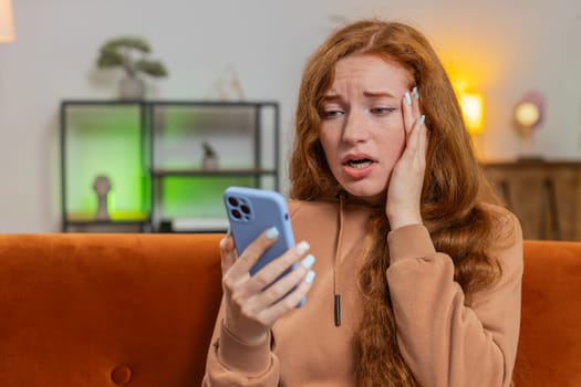 Upset displeased Caucasian woman using smartphone sitting on sofa couch in living room. Young adult redhead girl in casual clothes loses becoming surprised by sudden lottery results or bad news.