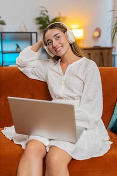 Happy overjoyed Caucasian woman in wireless headphones sitting on home sofa choosing listening disco music while working on laptop. Girl in white dress enjoy weekend activities entertaining. Vertical