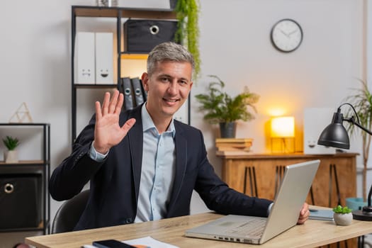 Hello. Happy young businessman employee waves hand hi gesture greeting welcomes someone, online webinar, remote distance conversation. Freelancer guy in suit sitting at desk with laptop in home office