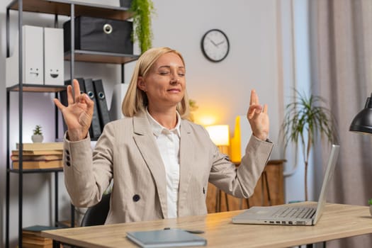 Caucasian mature businesswoman working on laptop, meditating doing yoga breathing exercise in lotus position at home office. Calm serene freelancer girl taking break. Peace of mind. Busy occupation.