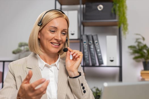 Confident Caucasian mature businesswoman in headset talking on video call using laptop at home office desk. Female advisor has online consultation, helps customers, customer support with complaints.