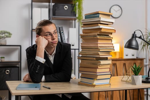 Tired exhausted stressed young businessman looking at large pile of books and looking at volume of work with sad face while preparing for exams at home office. Overwork education business concept.