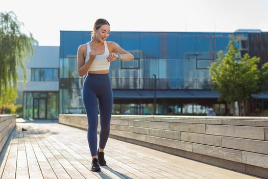Athletic runner woman looks at smartwatch device fitness bracelet checking time, heart rate, pulse, SPO2 on smartwatch while jogging. Young girl working out cardio outside in sunny urban city park.