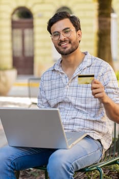 Indian man using credit bank card laptop computer while transferring money, purchases online shopping, order food delivery booking hotel room outdoors. Young guy tourist sitting on chair in city.