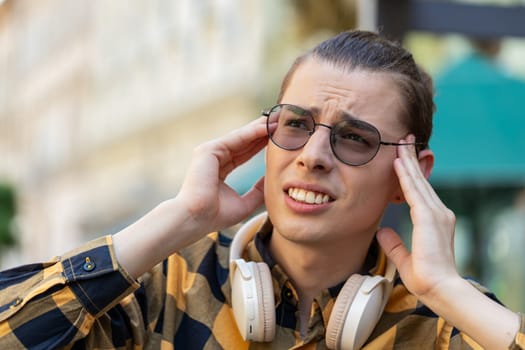 Displeased exhausted Caucasian man male rubbing temples to cure headache problem suffering from tension migraine stress. Young guy tourist in sunglasses grimacing, pain standing on city urban street.