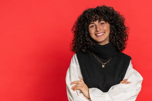 Portrait of happy Caucasian young woman with curly hair smiling friendly, glad expression looking at camera dreaming, resting, relaxation feel satisfied good news. Girl indoors on red background
