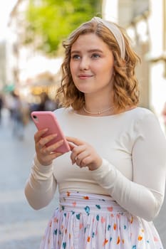 Redhead woman in dress using smartphone typing texting social media messages e-mail looking for a way on map in mobile navigator app outdoors. Girl walking in city street. Town lifestyles. Vertical
