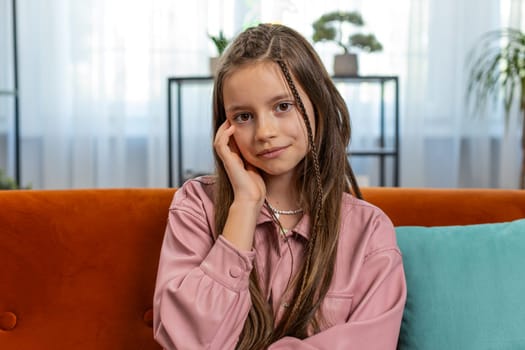 Portrait of happy smiling young girl child sitting on comfy sofa couch looking at camera in living room at home apartment. Cute teenage female school kid with brown long hair. Lifestyles.