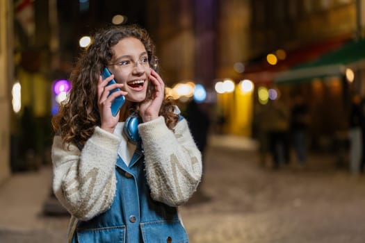 Phone call, good news, gossip. Happy excited Caucasian teenage girl in pleasant conversation on smartphone, enjoying talking with friend outdoors. Tourist in city street at night. Town lifestyles.