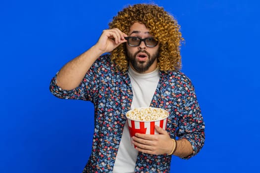 Excited man with curly hairstyle wig in 3D glasses eating popcorn and watching interesting tv serial, sport game, cinema film online social media movie content. Stylish guy isolated on blue background