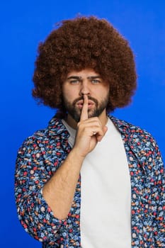 Shh be quiet please. Caucasian man presses index finger to lips makes silence gesture sign do not tells secret, stop talk gossip, confidential privacy. Stylish young guy isolated on blue background