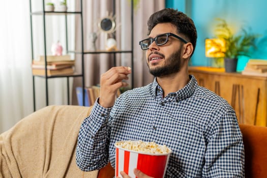 Happy Indian man taking break from work wearing 3D glasses eating popcorn watching movie film content on TV. Smiling Hispanic guy sits at home couch sofa having snacks during weekends. Lifestyle