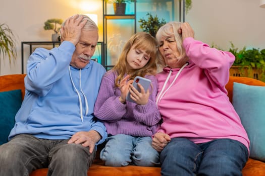 Upset grandfather, grandmother and granddaughter playing game on smartphone and losing at home. Sad Caucasian girl with grandparents received bad news, fortune loss, fail education study test on sofa.