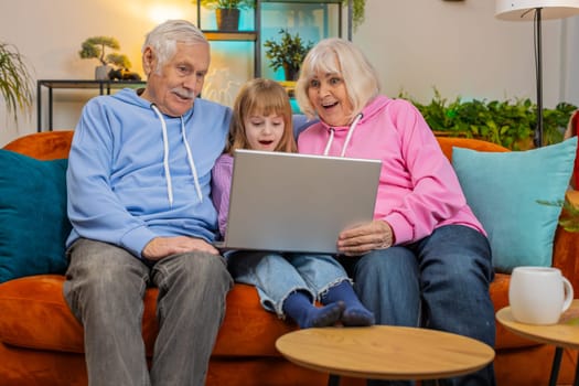 Elderly Caucasian couple man and woman with child girl granddaughter spending time at home together, sitting on sofa in the living room, using laptop computer. Watching videos playing games on weekend