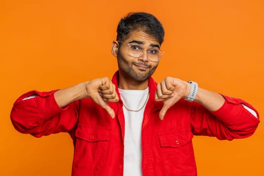 Dislike. Upset unhappy Indian man showing thumbs down sign gesture, expressing discontent, disapproval, dissatisfied negative feedback. Attractive bearded guy indoors isolated on orange background