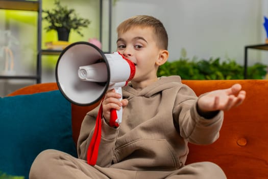 Preteen child boy talking with megaphone, proclaiming news, loudly announcing advertisement warning using loudspeaker to shout speech. Teenager male little kid at home play room apartment on couch