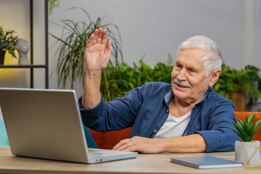 Senior man making video webcam conference call with friends or family, waving hello, enjoying pleasant conversation at home apartment indoors. Elderly mature grandfather sits on couch at table desk