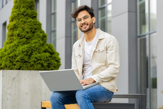 Happy young Indian man freelancer working on laptop computer sends messages, makes online purchases, watching movies, reading email outdoors. Arabian Hindu guy sitting on street bench. Business people