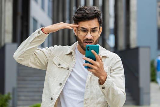 Surprised Indian man use smartphone typing browsing, loses becoming surprised sudden lottery results, bad news, fortune loss, fail outdoors. Arabian Hindu guy in downtown city street. Business people
