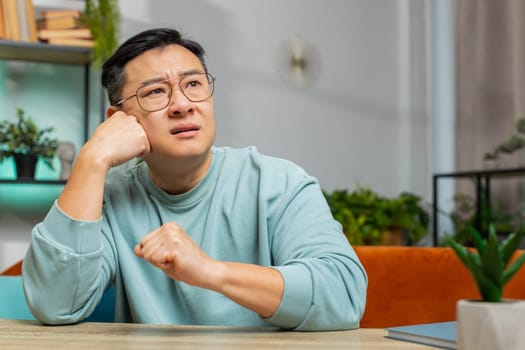 Portrait of sad Asian man guy sitting at home desk looks pensive thinks over life concerns or unrequited love, suffers from unfair situation. Problem, break up, depressed feeling bad annoyed, burnout