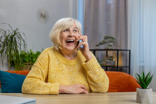 Phone call, good news, gossip. Happy smiling senior woman in pleasant conversation on smartphone, enjoying talking with friend at home apartment. Elderly mature old grandmother sits on couch at table