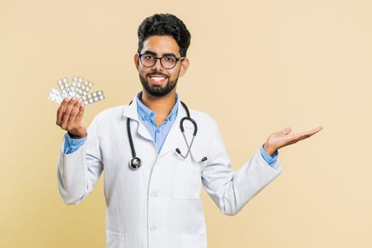 Indian doctor cardiologist man shows lot of tablet pills in blisters, recommends drugs vitamin antibiotic cure treatment. Health care. Arabian apothecary pharmacist guy isolated on beige background