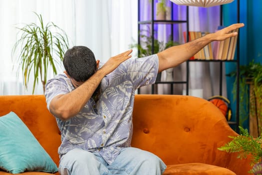 Trendy cheerful positive Indian Arabian man having fun dancing, dabbing raising hands making dub winner dance gesture celebrating victory at home apartment. Happy bearded Indian guy in room on sofa