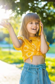 Happy preteen child kid having remote conversation communicate speaking by smartphone. Pretty girl daughter talking with mother on phone good news gossip standing on city sunset park street. Vertical